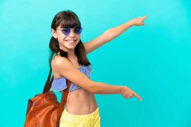 Küçük beyaz bir çocuk kumsala gidiyor. Mavi arka planda izole edilmiş bir şekilde. Parmağıyla yan tarafı gösteriyor ve bir ürün sunuyor.