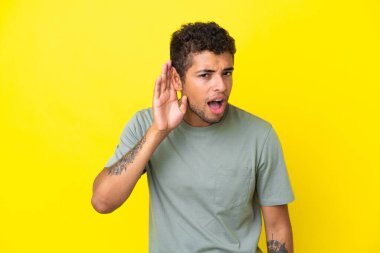 Genç ve yakışıklı Brezilyalı bir adam sarı arka planda izole edilmiş bir şekilde elini kulağına koyarak bir şeyler dinliyor.