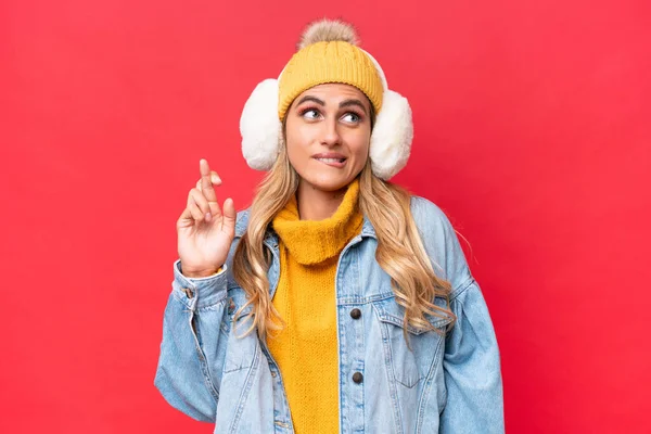 Young pretty Uruguayan woman wearing winter muffs isolated on red background background with fingers crossing and wishing the best