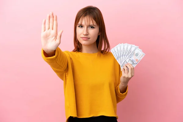 stock image Redhead girl taking a lot of money isolated on pink background making stop gesture