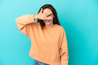 Mavi arka planda izole edilmiş genç beyaz bir kadın. Gözleri elleriyle kaplıyor ve gülümsüyor.