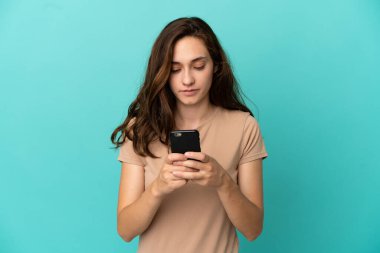 Genç beyaz kadın mavi arka planda cep telefonu kullanıyor.