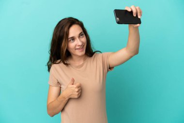 Mavi arka planda izole edilmiş beyaz bir kadın cep telefonuyla selfie çekiyor.