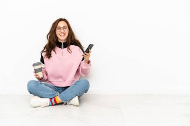 Beyaz arka planda izole bir şekilde yerde oturan beyaz tenli genç bir kadın kahve alıp götürmek için elinde bir cep telefonu...