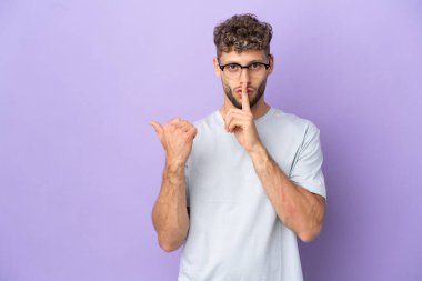 Mor arka planda izole edilmiş beyaz bir adam var. Yan tarafı işaret ediyor ve sessizlik hareketi yapıyor.