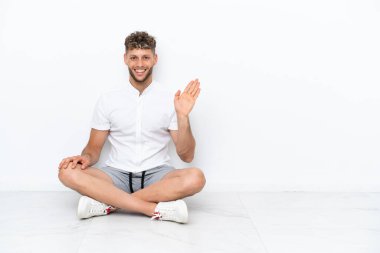 Genç sarışın adam, beyaz arka planda izole bir şekilde yerde oturuyor. Mutlu bir ifadeyle selamlıyor.