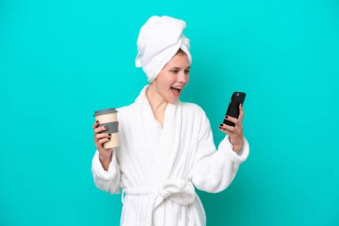 Bornozlu genç bir kadın mavi arka planda izole edilmiş. Elinde kahve ve cep telefonu var.