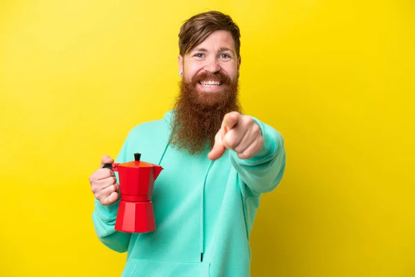 Redhead man with beard holding coffee pot isolated on yellow background points finger at you with a confident expression