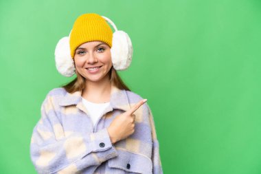 İzole edilmiş krom arka plan üzerine kış maskesi takan genç sarışın kadın bir ürünü sunmak için kenarı işaret ediyor.