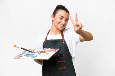 İzole edilmiş beyaz arka planda palet tutan genç sanatçı gülümsüyor ve zafer işareti gösteriyor.