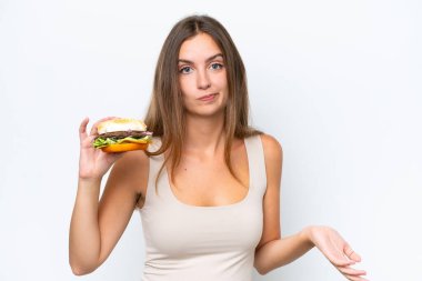 Genç, güzel bir kadın, beyaz arka planda izole edilmiş bir burger tutuyor. Omuzlarını kaldırırken şüphe uyandırıyor.