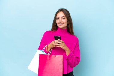 Genç ve güzel bir kadın mavi arka planda izole edilmiş. Elinde alışveriş çantaları var ve bir arkadaşına cep telefonuyla bir mesaj yazıyor.
