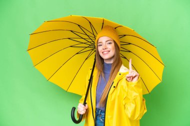 Yağmura dayanıklı ceketi ve şemsiyesi olan genç ve güzel bir kadın izole edilmiş krom anahtar arka planında en iyisini gösteren ve kaldıran parmağını kaldıran
