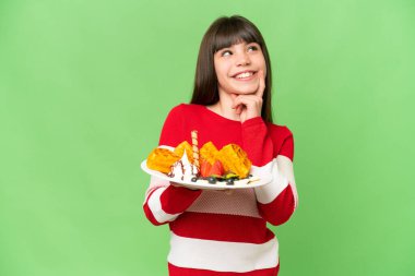 İzole edilmiş krom arka planda waffle tutan küçük kız yukarı bakarken bir fikir düşünüyor.