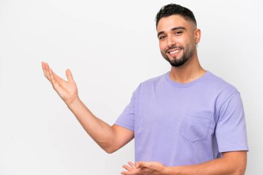 Beyaz arka planda soyutlanmış genç Arap yakışıklı adam davet edilmek için ellerini yana uzatıyor.