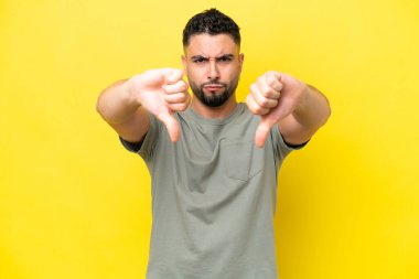 Sarı arka planda iki eliyle baş parmağını gösteren yakışıklı genç bir Arap.