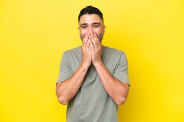 Genç Arap yakışıklı adam sarı arka planda izole edilmiş mutlu ve gülümseyen elleriyle ağzını kapatıyor.