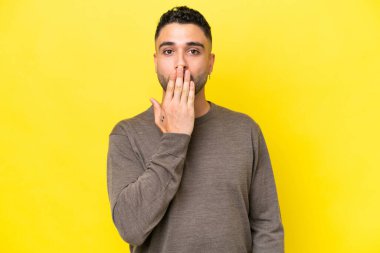 Sarı arka planda izole edilmiş genç Arap yakışıklı adam ağzını eliyle kapatıyor.