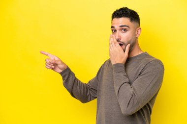 Sarı arka planda yalıtılmış genç Arap yakışıklı bir adam sürpriz bir ifadeyle işaret ederken