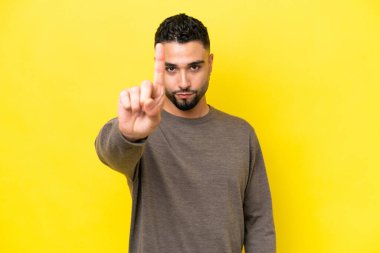 Sarı arka planda izole genç Arap yakışıklı adam ciddi bir ifadeye sahip birini sayıyor.