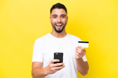 Genç Arap yakışıklı adam sarı arka planda izole edilmiş cep telefonuyla alışveriş yapıyor ve elinde şaşırmış bir ifadeyle bir kredi kartı tutuyor.