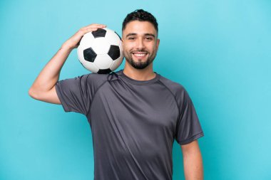 Genç Arap yakışıklı adam mavi arka planda futbol topuyla izole edilmiş.