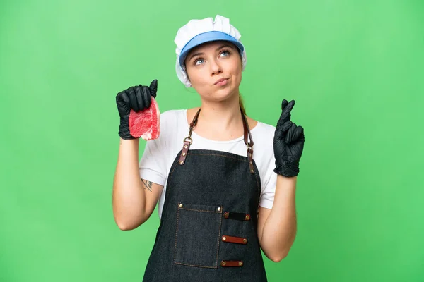 Butcher woman wearing an apron and serving fresh cut meat over isolated chroma key background with fingers crossing and wishing the best