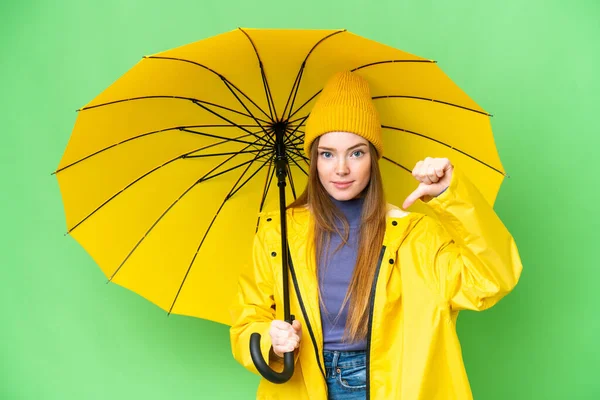 年轻漂亮的女人 穿着防雨外套 带着雨伞 罩在孤立的彩色关键背景上 带着负面表情垂下大拇指 — 图库照片