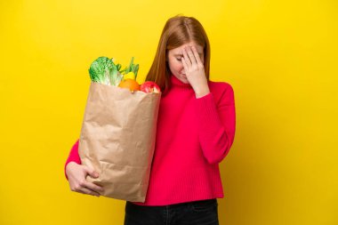 Sarı arka planda izole edilmiş alışveriş torbası tutan genç kızıl saçlı kadın yorgun ve hasta bir ifadeyle.