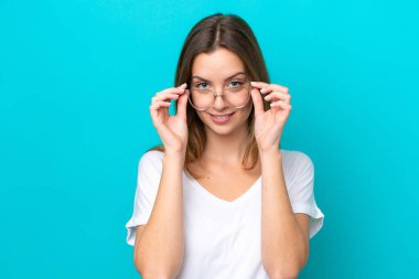 Genç beyaz kadın mavi arka planda izole edilmiş, mutlu bir ifadeyle gözlüklü.