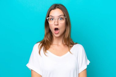 Mavi arka planda gözlüklü ve şaşkın bakışlı genç beyaz kadın.