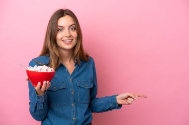 Beyaz bir kadın elinde bir kase mısır gevreği tutuyor pembe arka planda izole edilmiş şaşırmış ve parmağıyla kenarı işaret ediyor.