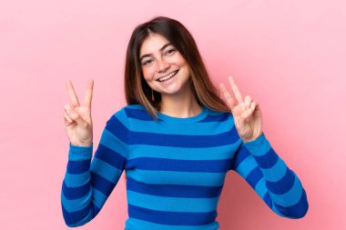 Pembe arka planda izole edilmiş genç beyaz kadın iki eliyle zafer işareti gösteriyor.