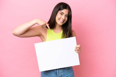 Pembe arka planda izole edilmiş genç Brezilyalı bir kadın elinde mutlu bir ifadeyle boş bir levha tutuyor ve onu işaret ediyor.