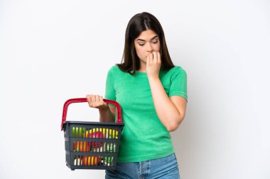Young Brazilian woman holding a shopping basket full of food isolated on white background having doubts