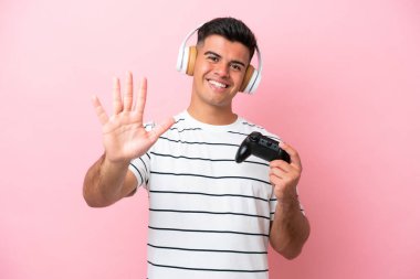 Young handsome man playing with a video game controller isolated on pink background counting five with fingers
