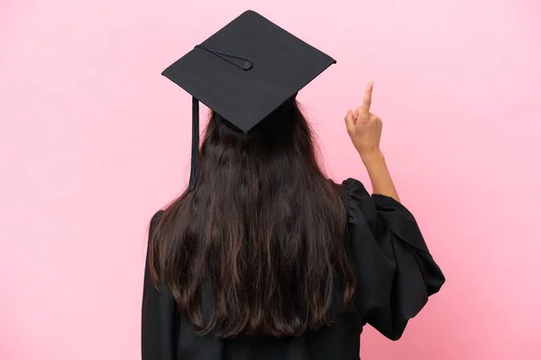 Young university Colombian woman graduate isolated on pink background pointing back with the index finger