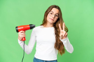 Teenager girl holding a hairdryer over isolated chroma key background smiling and showing victory sign