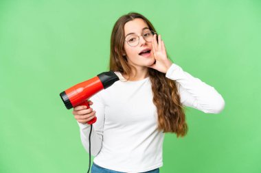 Teenager girl holding a hairdryer over isolated chroma key background shouting with mouth wide open