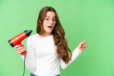 Teenager girl holding a hairdryer over isolated chroma key background surprised and pointing finger to the side