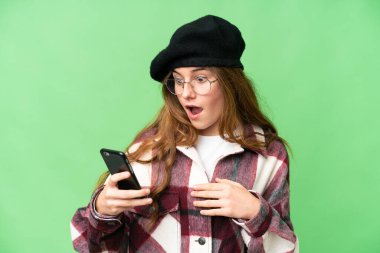 Teenager girl over isolated chroma key background looking at the camera while using the mobile with surprised expression