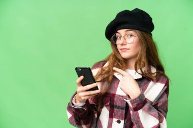 Teenager girl over isolated chroma key background using mobile phone and pointing back