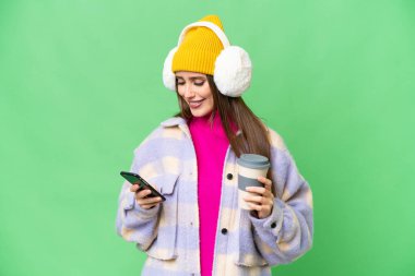 Young woman wearing winter muffs over isolated chroma key background holding coffee to take away and a mobile