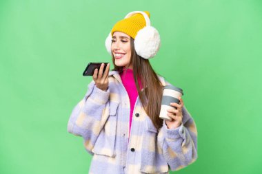 Young woman wearing winter muffs over isolated chroma key background holding coffee to take away and a mobile
