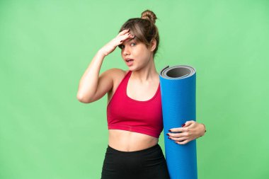 Genç sporcu kız yoga derslerine gidiyor. Elinde paspas, izole edilmiş krom anahtar arkaplanının üzerinde. Yan tarafa bakarken sürpriz jest yapıyor.