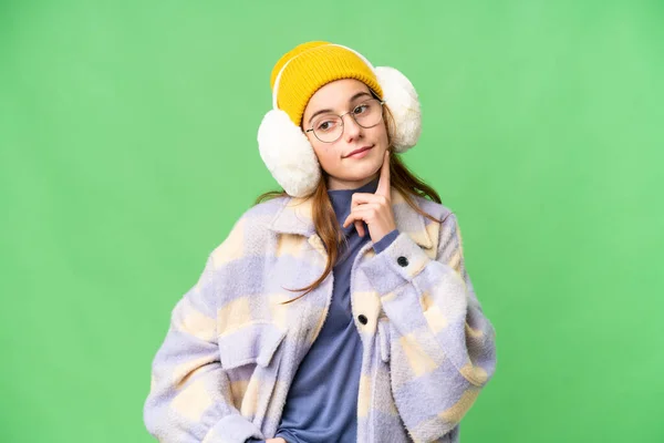 Teenager girl wearing winter muffs over isolated chroma key background thinking an idea while looking up