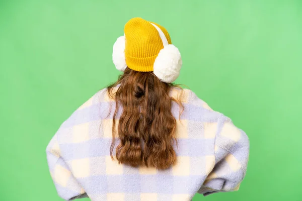 Teenager girl wearing winter muffs over isolated chroma key background in back position