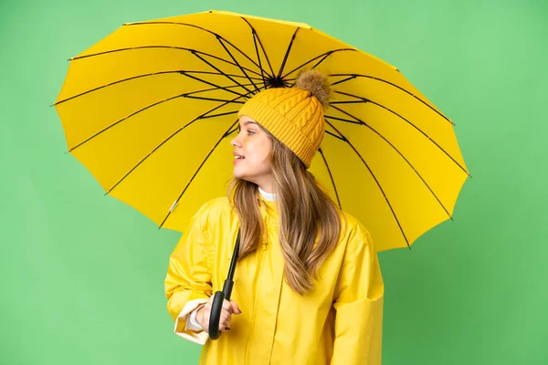 穿着防雨外套和雨伞的小女孩在孤零零的彩色关键背景上看一边 — 图库照片