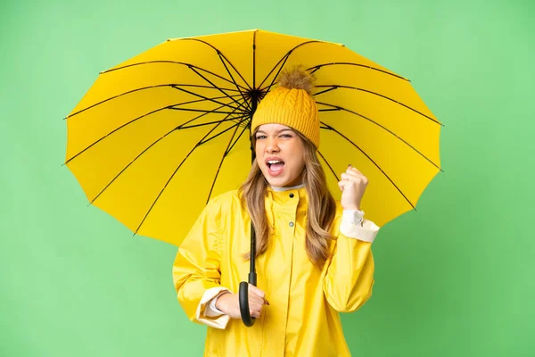 年轻姑娘身穿雨衣 披着雨伞 在孤零零的彩色关键背景下庆祝胜利 — 图库照片
