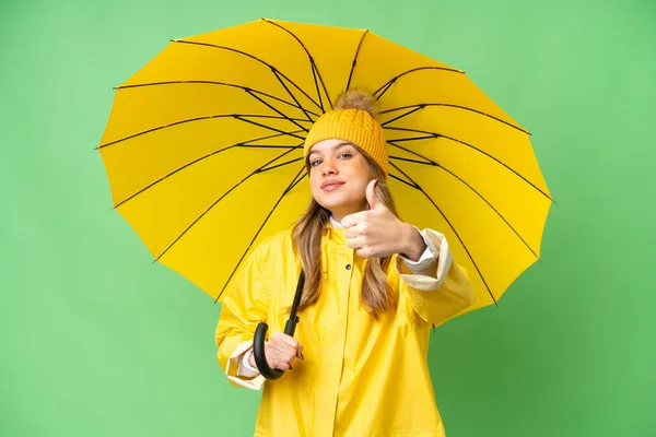 年轻姑娘穿着雨衣 披着雨伞 戴在孤立的彩色关键背景上 竖起大拇指 因为发生了件好事 — 图库照片